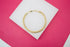 A polished gold cuban link bracelet rests in a circle on a white platform against a vibrant pink background.