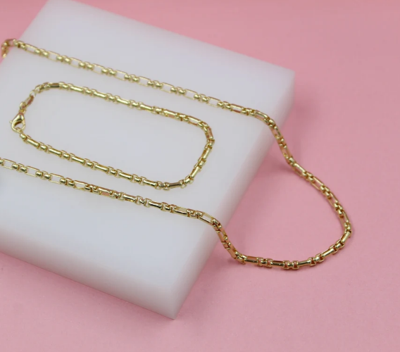 A matching gold barrel-link necklace and bracelet set rests on a white display block against a soft pink background.