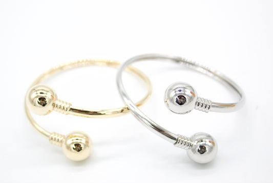 A gold cuff bracelet and a silver cuff bracelet rest side-by-side on a plain white surface, each featuring shiny spherical ball ends and textured ribbed detailing.
