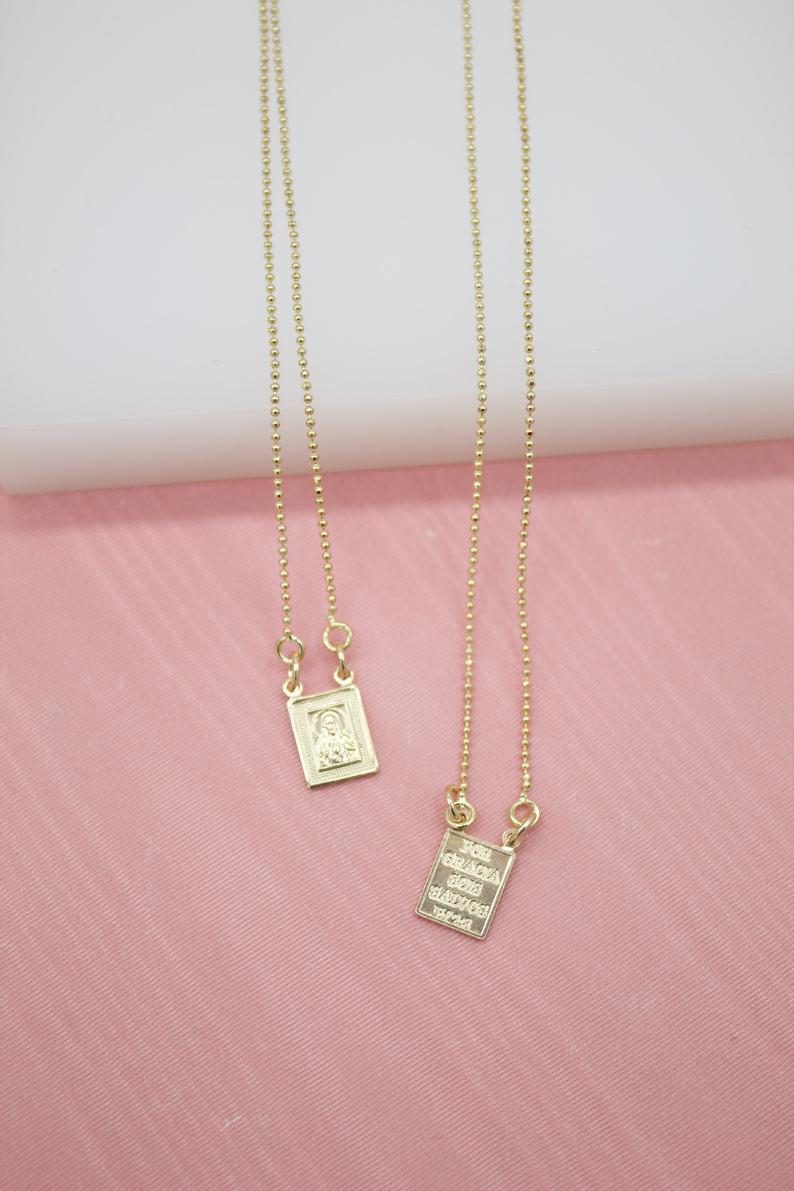 Two delicate gold ball-chain necklaces feature small rectangular religious pendants resting on a soft pink surface.
