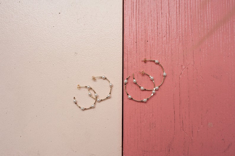 Several gold hoop earrings adorned with small white pearls rest on a split-color pink and cream wooden background.