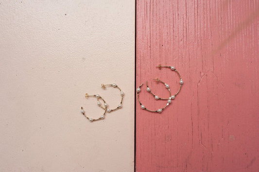 Several gold hoop earrings adorned with small white pearls rest on a split-color pink and cream wooden background.