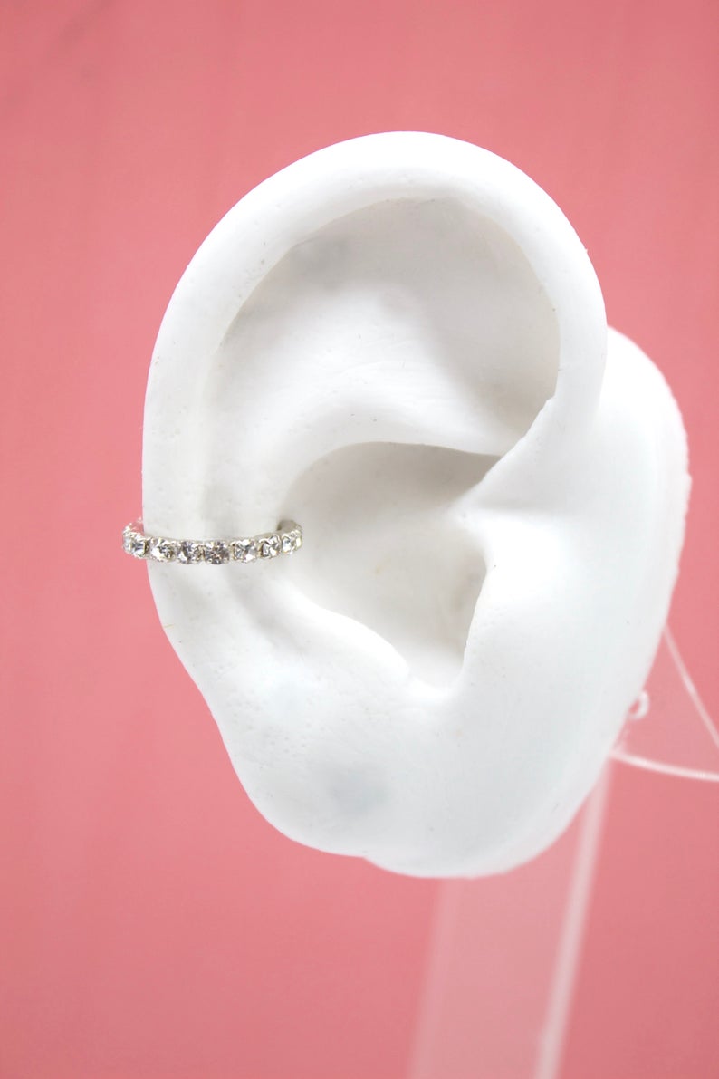 A white model ear displays a sparkling diamond cartilage hoop earring against a pink background.