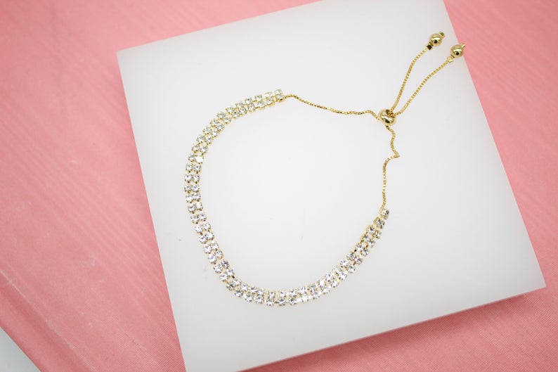 A gold tennis bracelet with double rows of sparkling rhinestones and an adjustable slider clasp rests on a white display surface against a pink background.