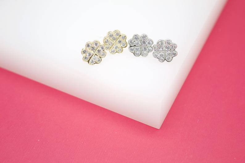 Four shimmering clover stud earrings featuring pave-set cubic zirconia crystals rest neatly on a white display surface, showcasing both gold and silver finishes.