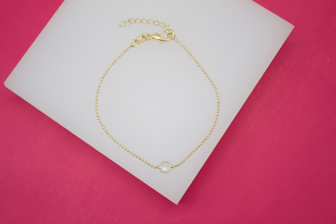 A delicate gold-plated bead chain bracelet featuring a single sparkling zircon crystal rests on a white display surface against a vibrant pink background.