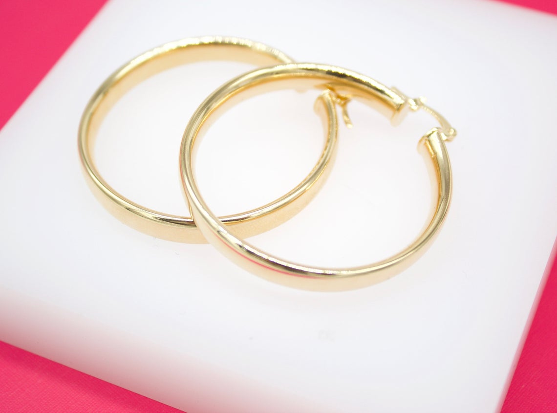A pair of polished gold hoop earrings rests elegantly on a white display surface against a vibrant pink background.