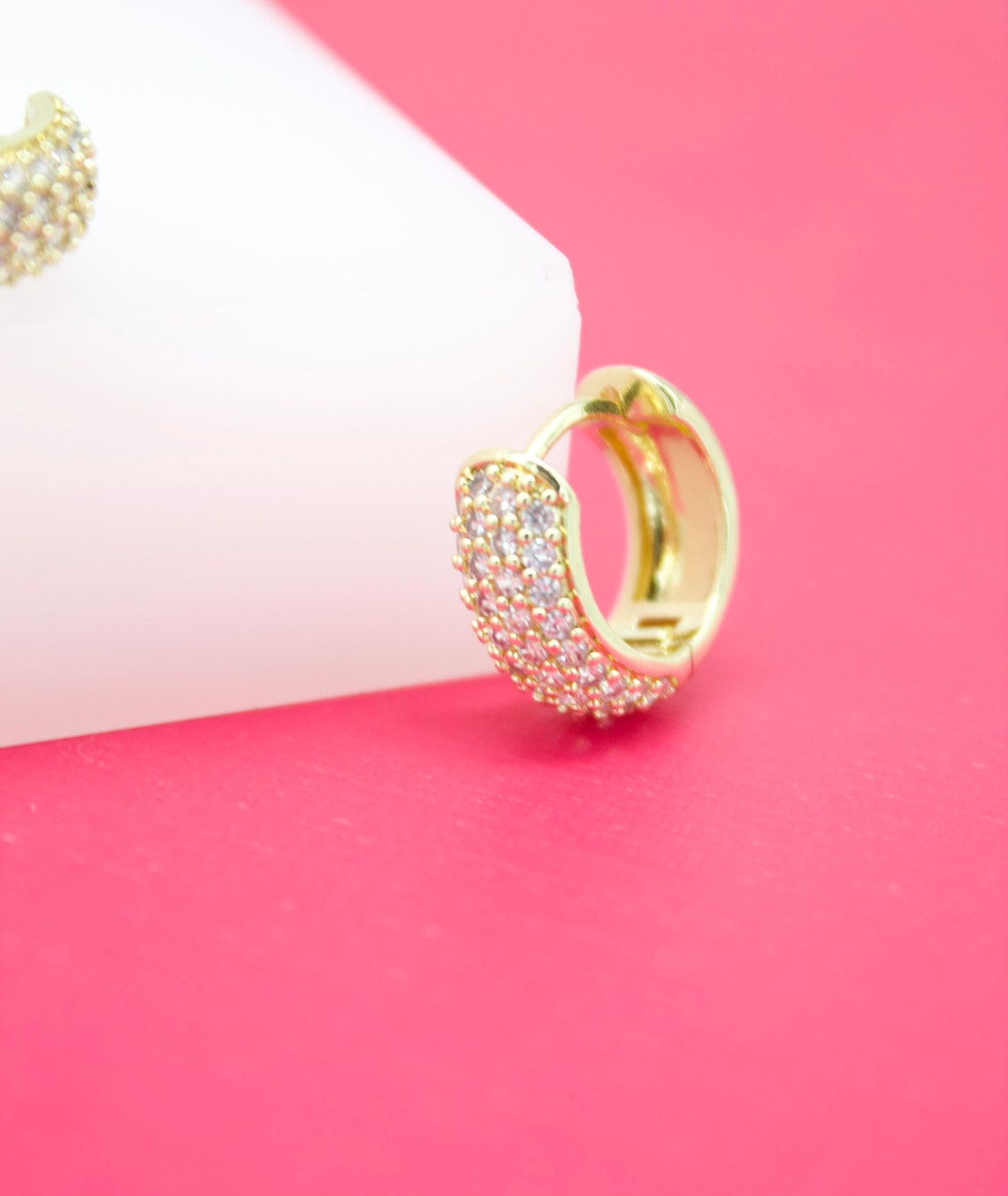 A sparkling gold huggie hoop earring encrusted with small pave crystals rests on a white block against a vibrant pink background.