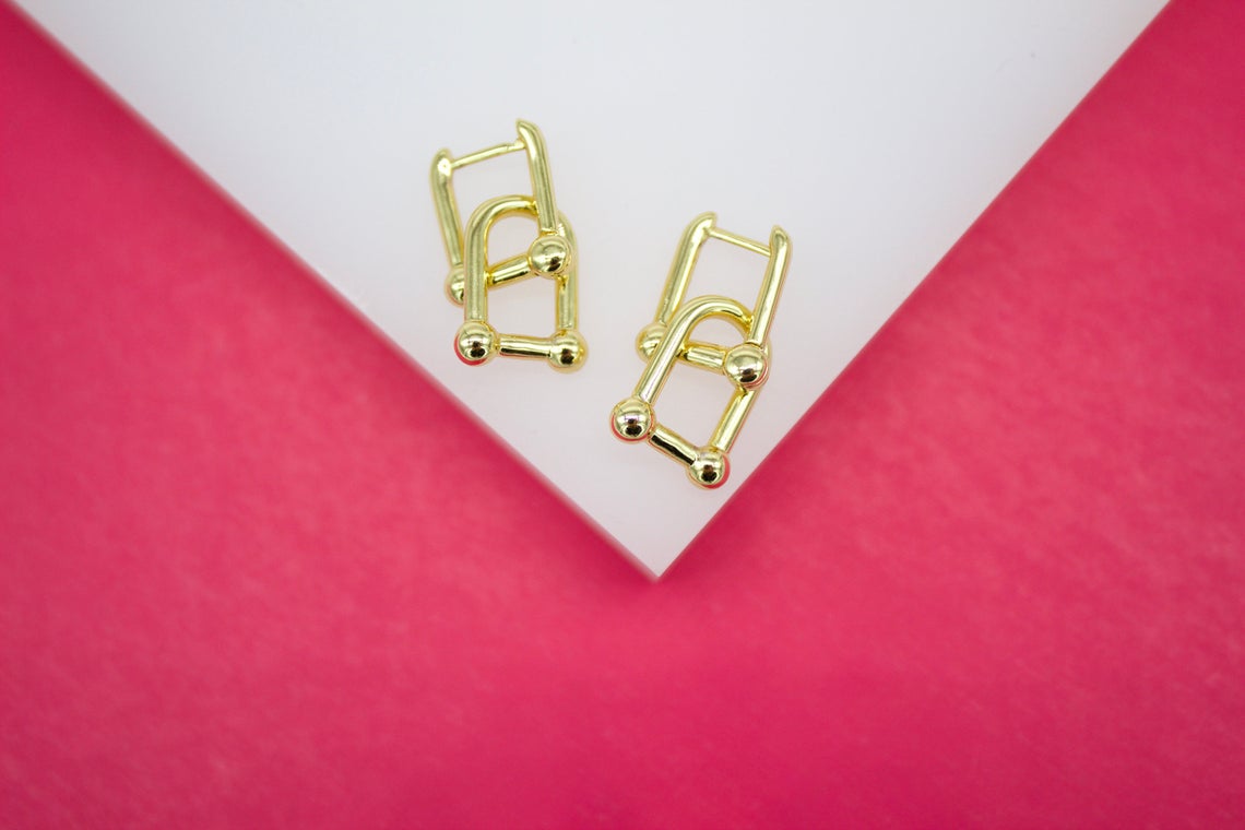A pair of geometric gold interlocking rectangular hoop earrings rests on a white display surface against a vibrant pink background.