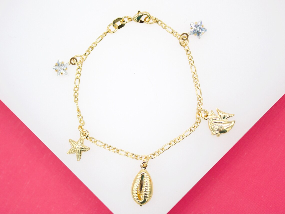 A gold charm bracelet features whimsical ocean-themed charms, including a seashell, starfish, fish, and sparkling crystal stars.