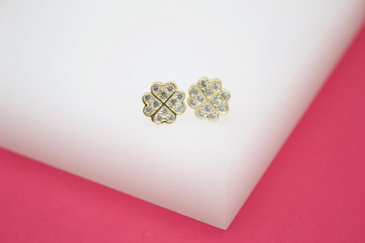 A pair of gold four-leaf clover stud earrings sparkle with embedded cubic zirconia stones on a white and pink surface.