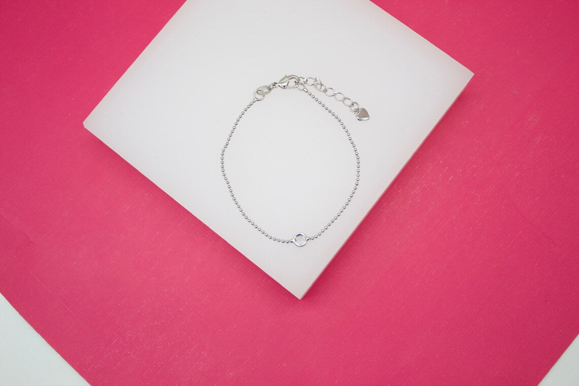 A delicate silver bead chain bracelet rests on a white display stand against a vibrant pink background.