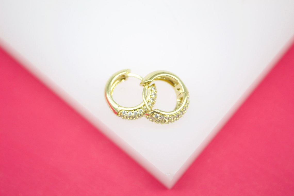 A pair of small gold hoop earrings encrusted with sparkling cubic zirconia stones rests on a white display stand against a vibrant pink background.