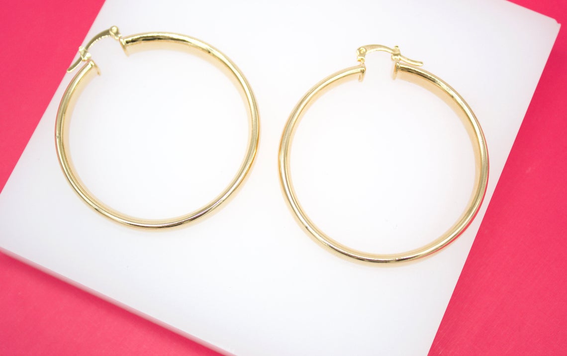 A pair of classic gold hoop earrings rests neatly on a white display stand against a vibrant pink background.