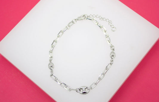A silver link bracelet featuring textured anchor chain elements rests on a white display block against a vibrant pink background.