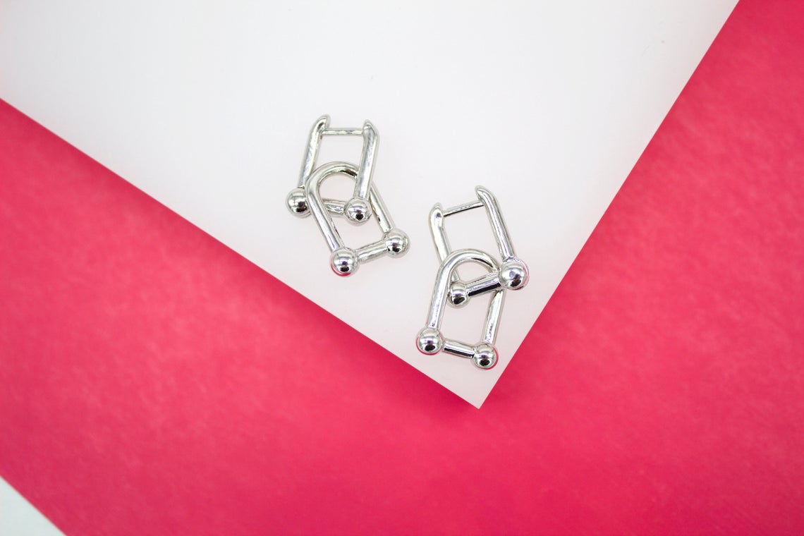 A pair of silver U-shaped hoop earrings featuring connected barbell links rests on a white surface against a bright pink background.