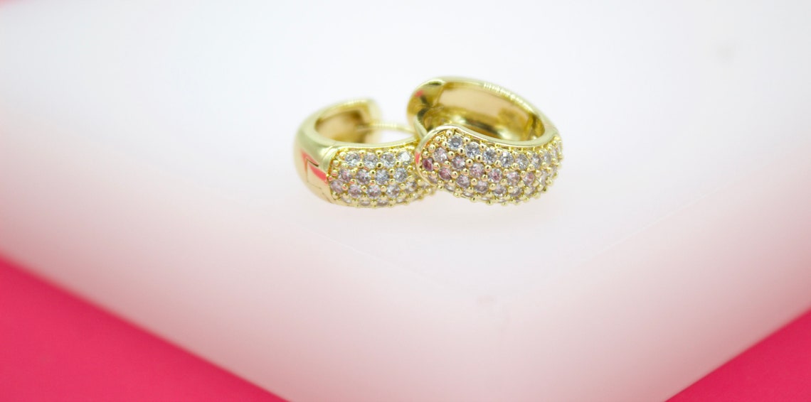 A pair of sparkling gold huggie hoop earrings encrusted with tiny pink and white cubic zirconia stones rests on a soft white surface.
