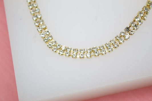 A gleaming double-strand rhinestone choker necklace rests on a white display surface against a soft pink background.