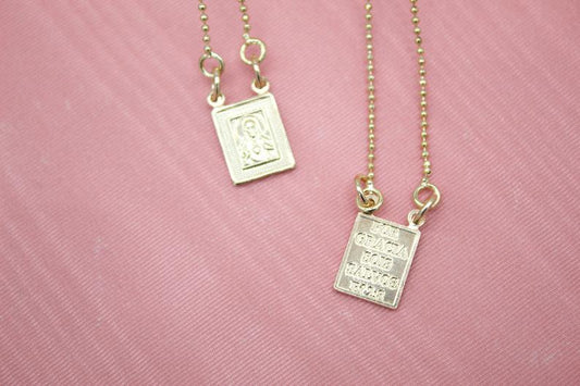 Two gold beaded necklaces with rectangular pendants rest against a pink fabric background, showcasing a religious figure on one pendant and engraved text on the other.