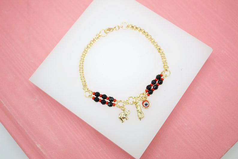 A gold chain bracelet adorned with black and red beads features dangling charms including an elephant, a figa, and an evil eye.