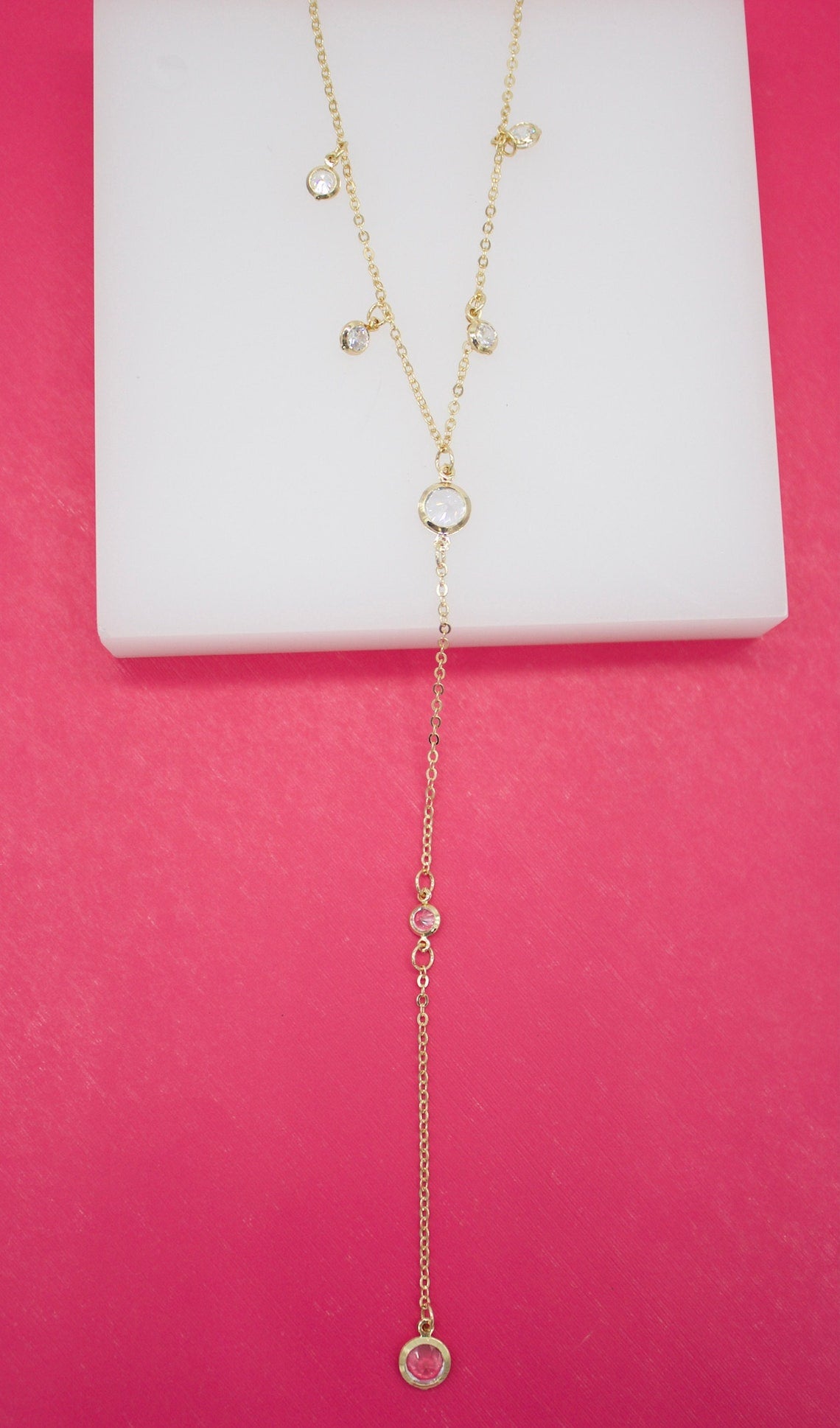 A delicate gold y-necklace featuring sparkling clear and pink gemstone charms rests on a white display against a vibrant pink background.
