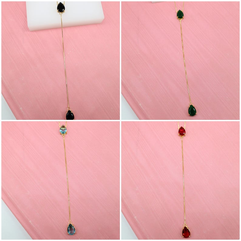 Four gold lariat necklaces feature teardrop-shaped gemstone pendants in black, green, blue, and red against a pink background.