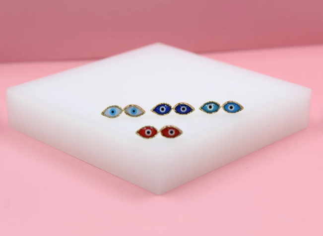 A collection of gold evil eye stud earrings in red, light blue, dark blue, and turquoise rest on a white block against a pink background.