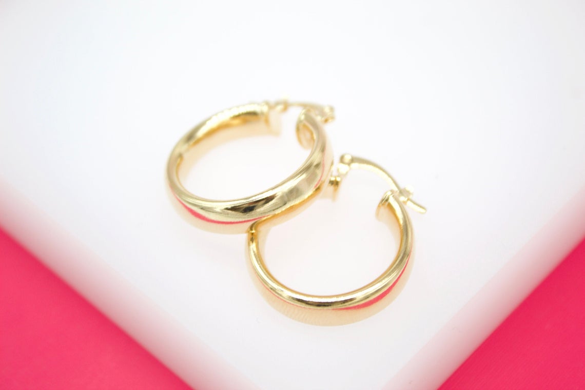 A pair of sleek gold hoop earrings rests on a clean white surface against a bright pink backdrop.