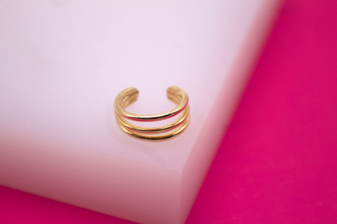 A gold triple-band ear cuff sits on a white display surface against a vibrant hot pink background.