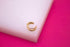 A polished triple-band gold ear cuff rests neatly on a white display block against a vibrant pink background.