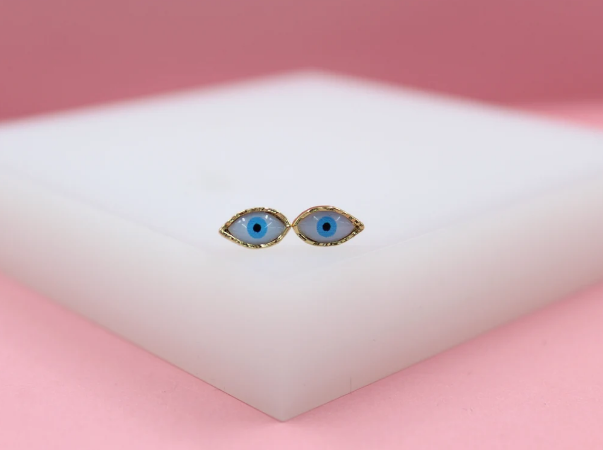 Two gold evil eye stud earrings with blue enamel rest on a white display block against a pink background.
