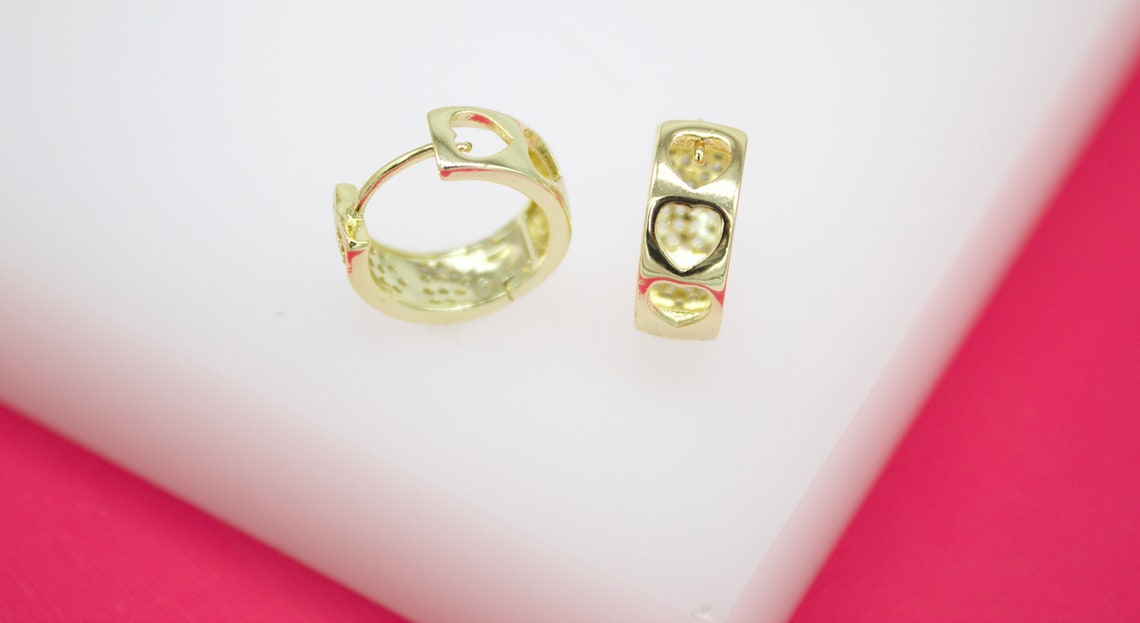 A pair of gold huggie hoop earrings featuring openwork heart designs rests on a white surface against a bright pink background.