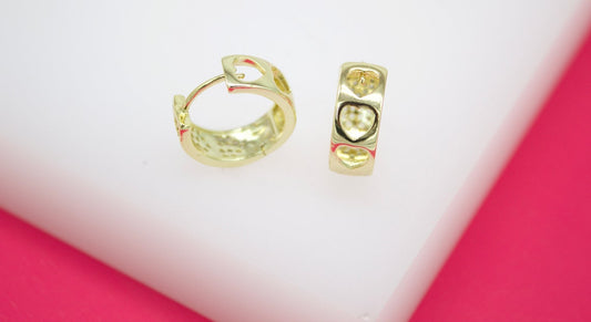 A pair of gold huggie hoop earrings featuring openwork heart designs rests on a white surface against a bright pink background.