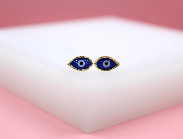 A pair of gold evil eye stud earrings rests on a white platform against a pink background.