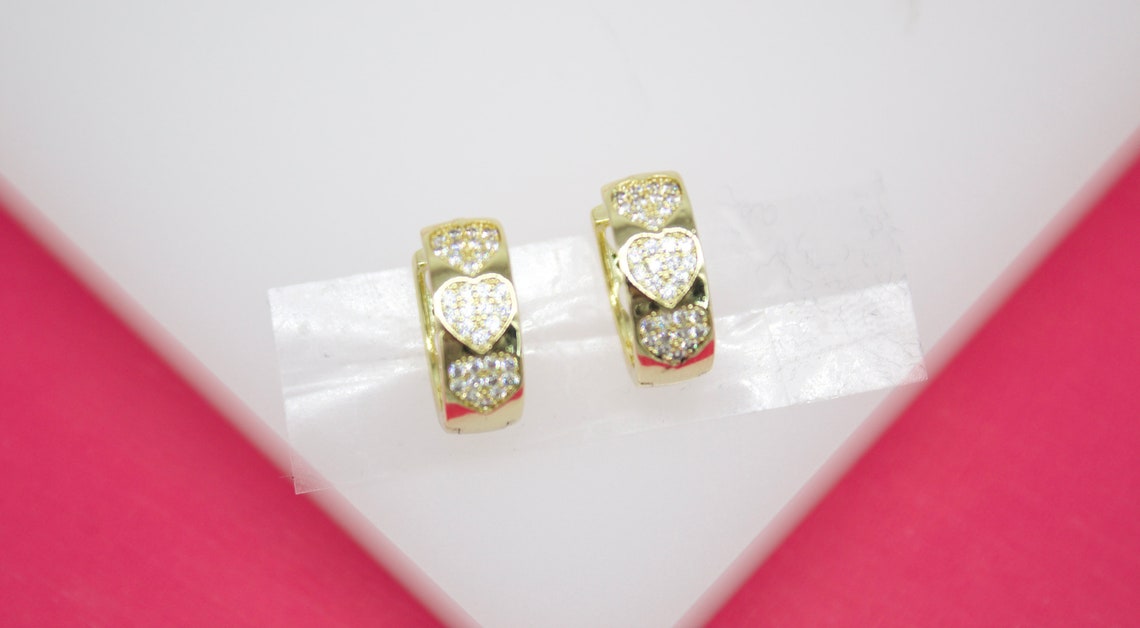 A pair of gold huggie hoop earrings decorated with sparkling pave-set heart designs rest on a white display stand against a pink background.