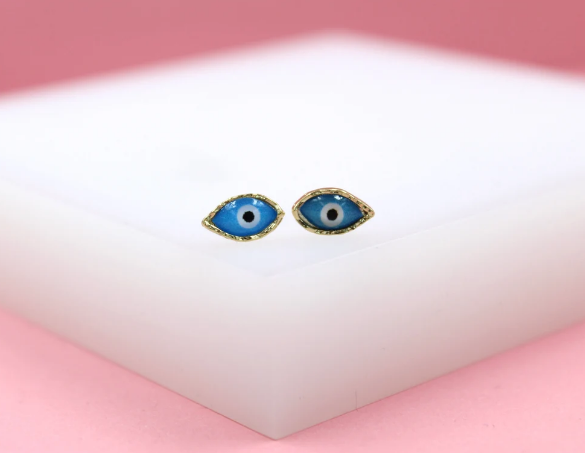 A pair of gold evil eye stud earrings featuring vibrant blue enamel rests on a white display against a pink backdrop.