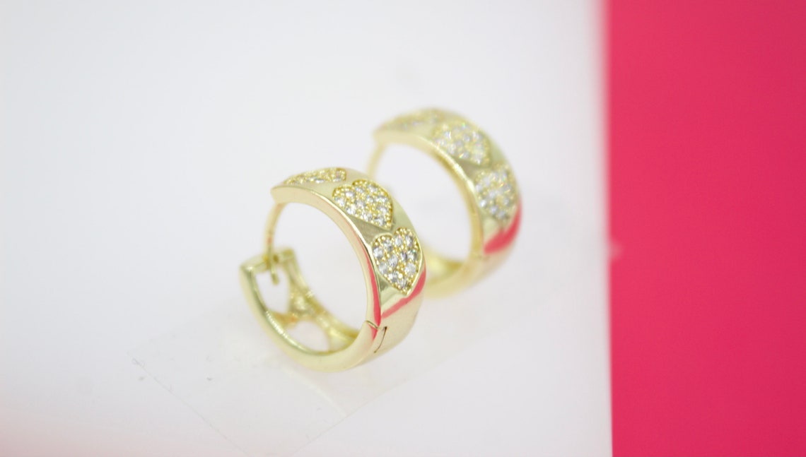 A pair of gold hoop earrings features sparkling pavé diamond hearts against a white and pink background.