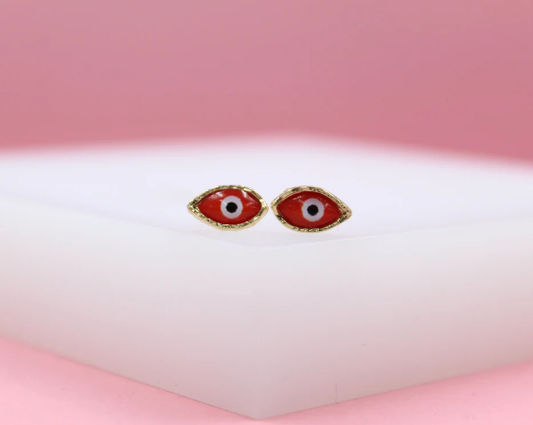 A pair of red evil eye stud earrings rest on a white display against a pink backdrop.
