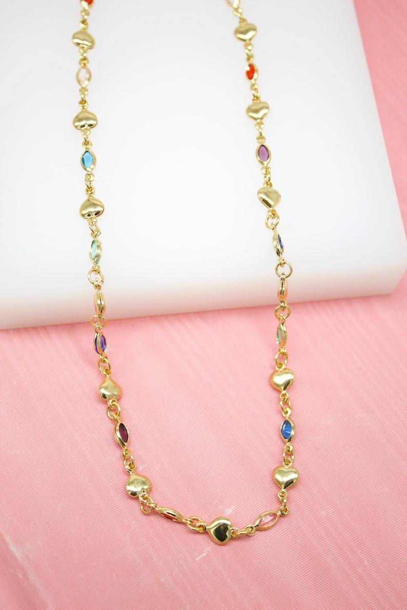 A gold chain necklace features alternating gold hearts and colorful gemstones against a pink and white background.
