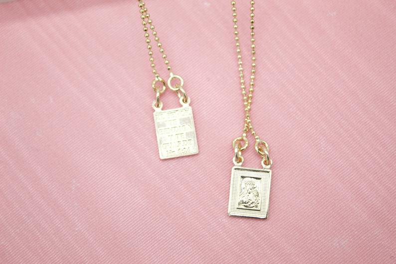 Two delicate gold necklaces with rectangular pendants featuring religious engravings rest against a soft pink background.