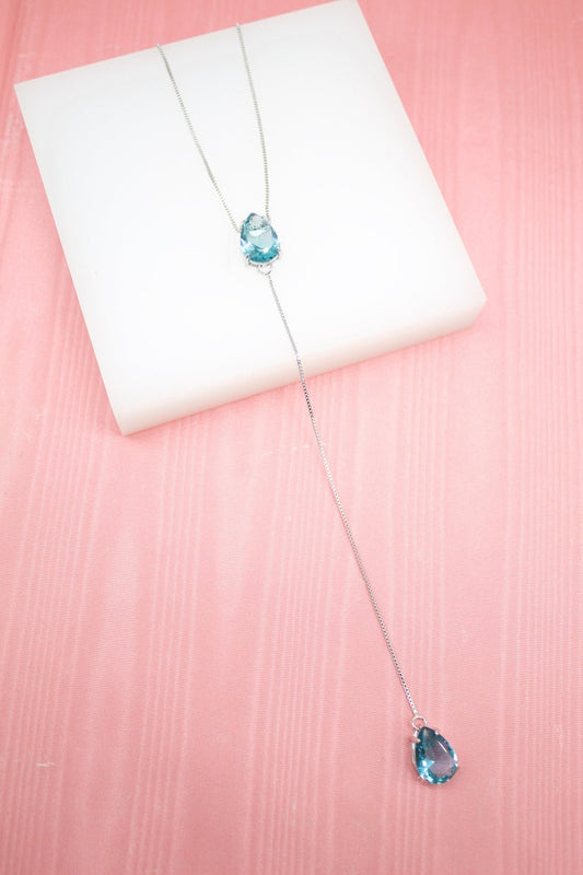 A delicate silver lariat necklace featuring two teardrop-shaped blue gemstones rests on a white display block against a pink textured background.