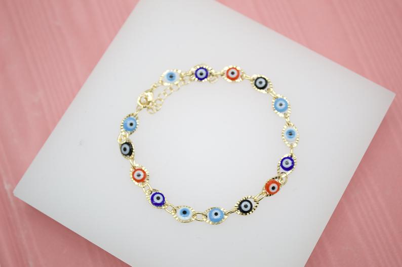 A delicate gold chain bracelet features multicolored evil eye charms resting on a white display card.