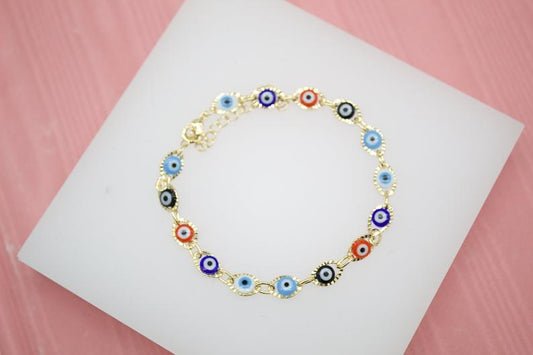 A delicate gold chain bracelet features multicolored evil eye charms resting on a white display card.