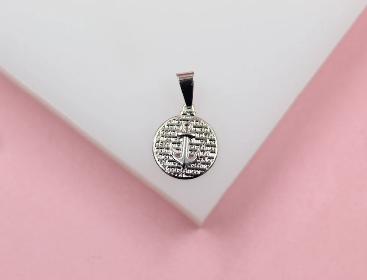 A silver anchor pendant with engraved script rests on a white display against a pink background.