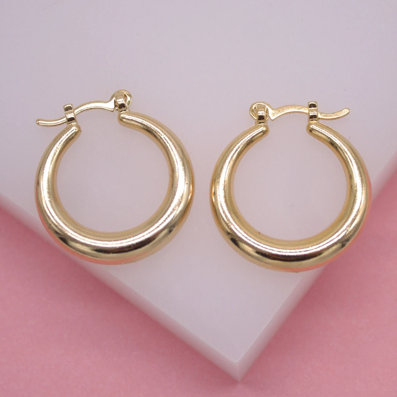 Two polished gold hoop earrings rest side by side on a soft white and pink surface.