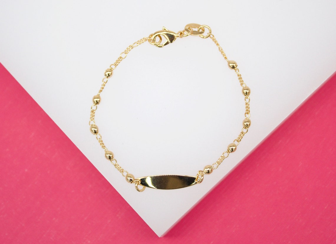 A delicate gold ID bracelet with interspersed beads lies against a pink and white background.