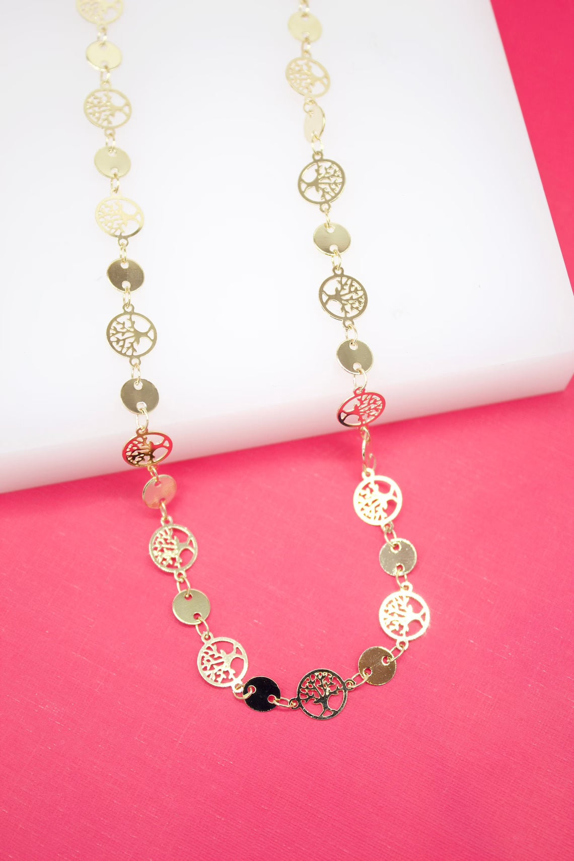 A long gold necklace features alternating coin links and intricate tree of life charms displayed against a vibrant pink and white background.