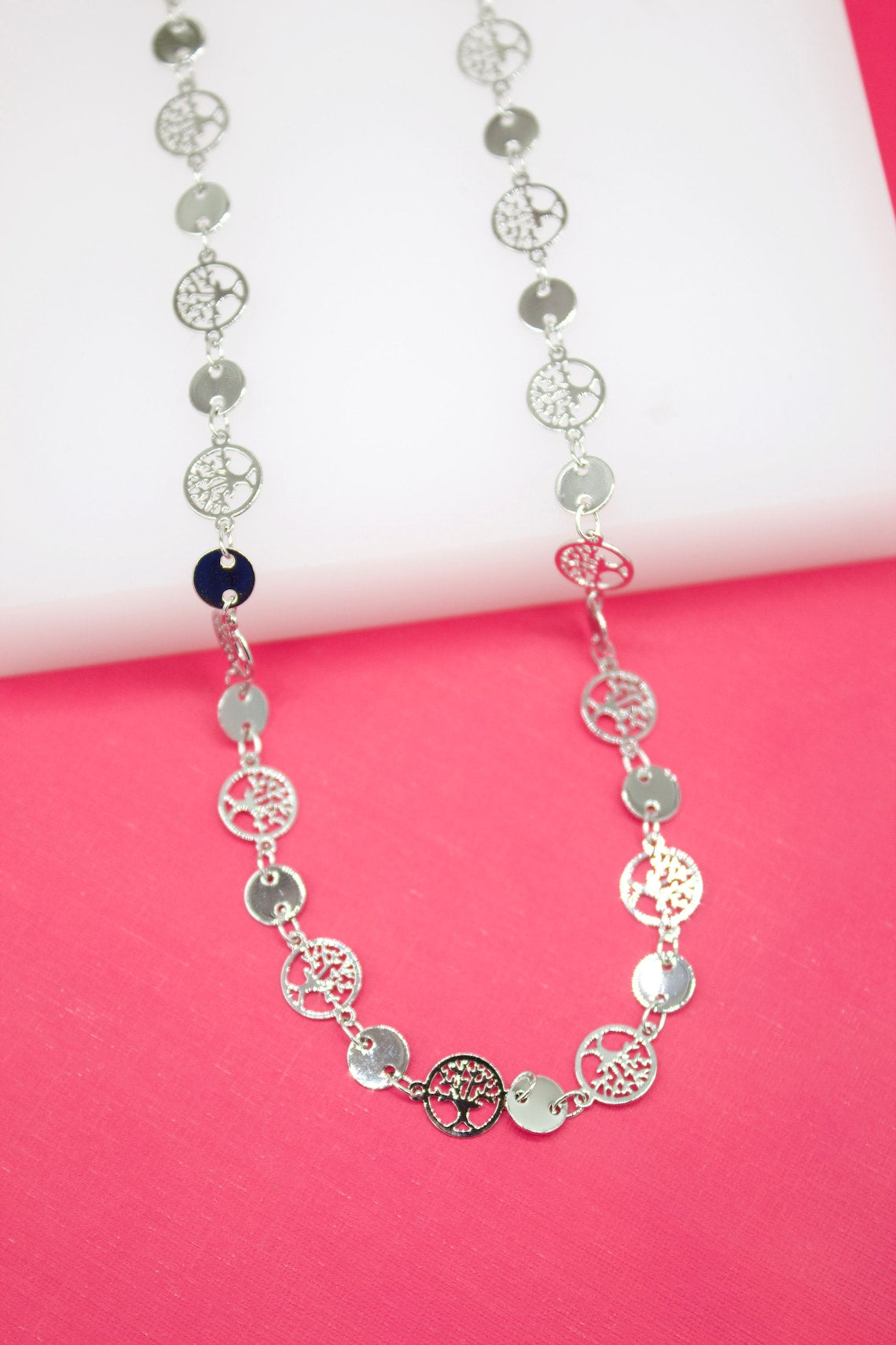 A long silver necklace featuring alternating smooth round discs and intricate tree of life pendants rests against a white and pink background.