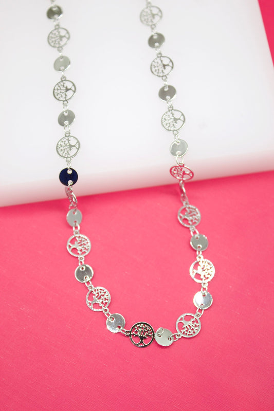 A delicate silver necklace features alternating smooth round disks and intricate tree of life charms displayed against a vibrant pink and white background.