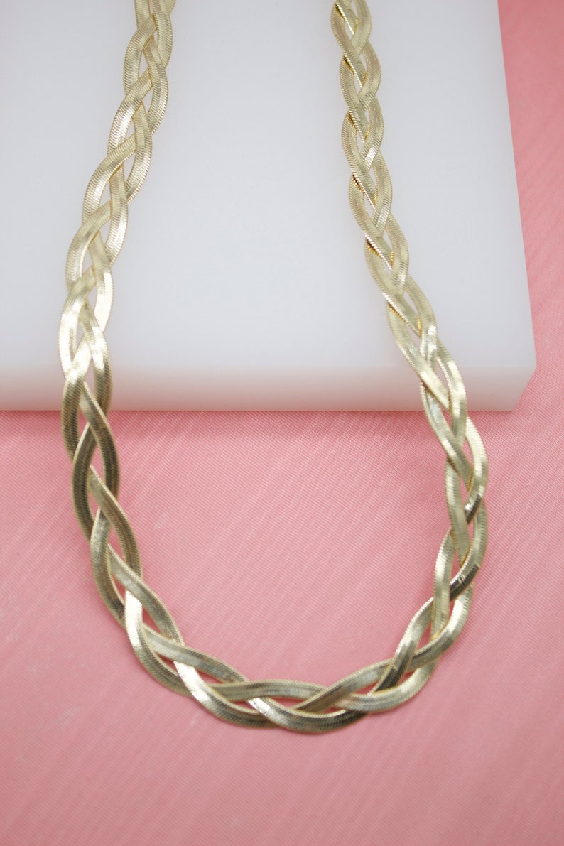 A gleaming gold braided herringbone chain necklace rests on a white display block against a pink fabric background.